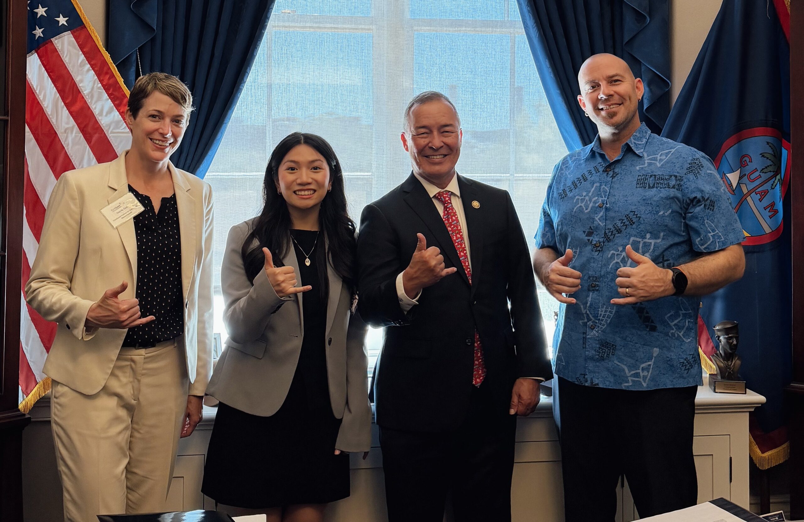 Four People Pose Together Indoors, Smiling and Giving Thumbs Up, with the American Flag and Guam Flag in the Background.