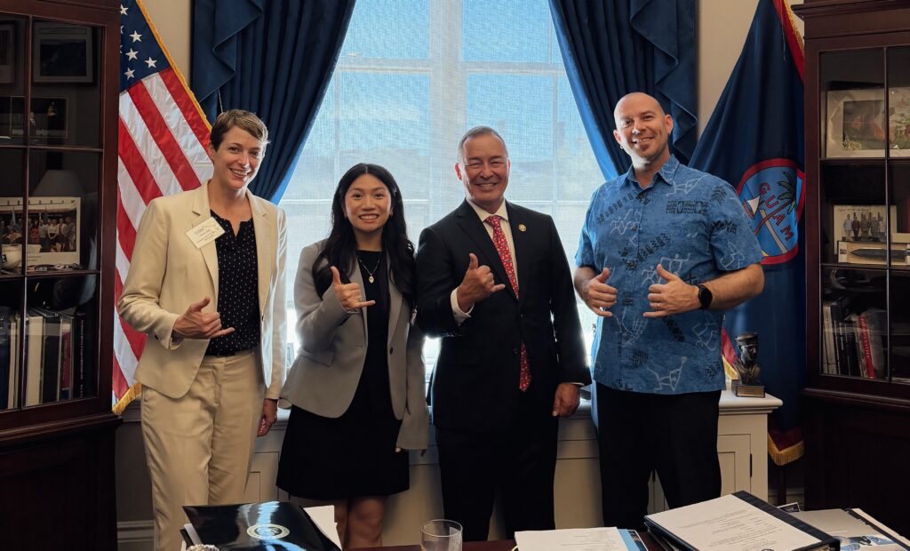Four Professionals Pose in a Formal Office, Giving Thumbs-up with U.s. and Guam Flags in the Background.