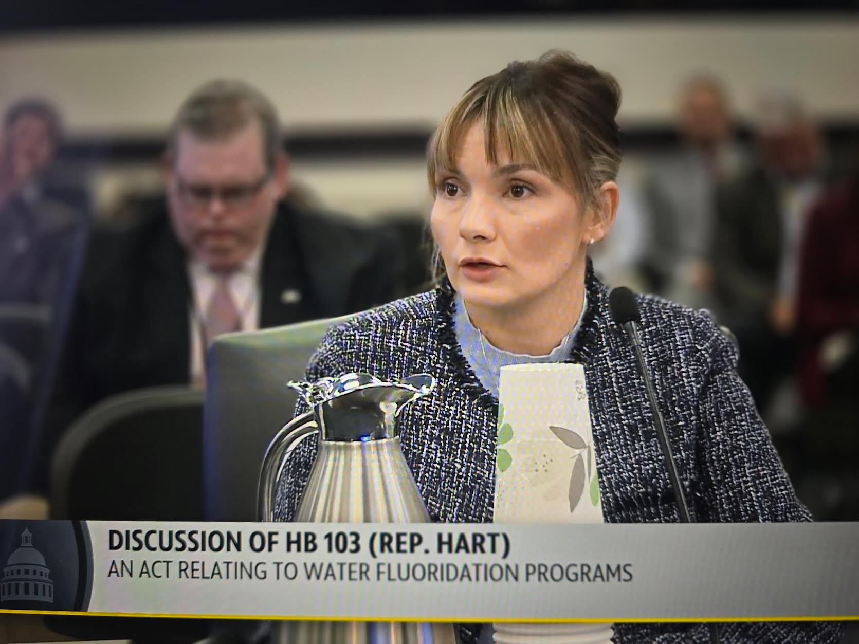 Woman Testifying at a Committee Hearing, Wearing a Tweed Jacket, with a Microphone and Documents in Front; Banner Reads 'Discussion of HB 103 (Rep. Hart)'.