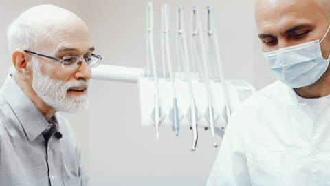Elderly Man with Glasses Speaks with a Dentist Wearing a Mask in a Dental Clinic, with Dental Instruments Visible in the Background.