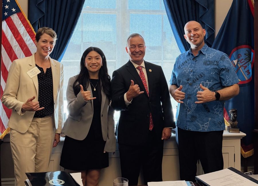 Four people pose together indoors, smiling and giving thumbs up, with the American flag and Guam flag in the background.