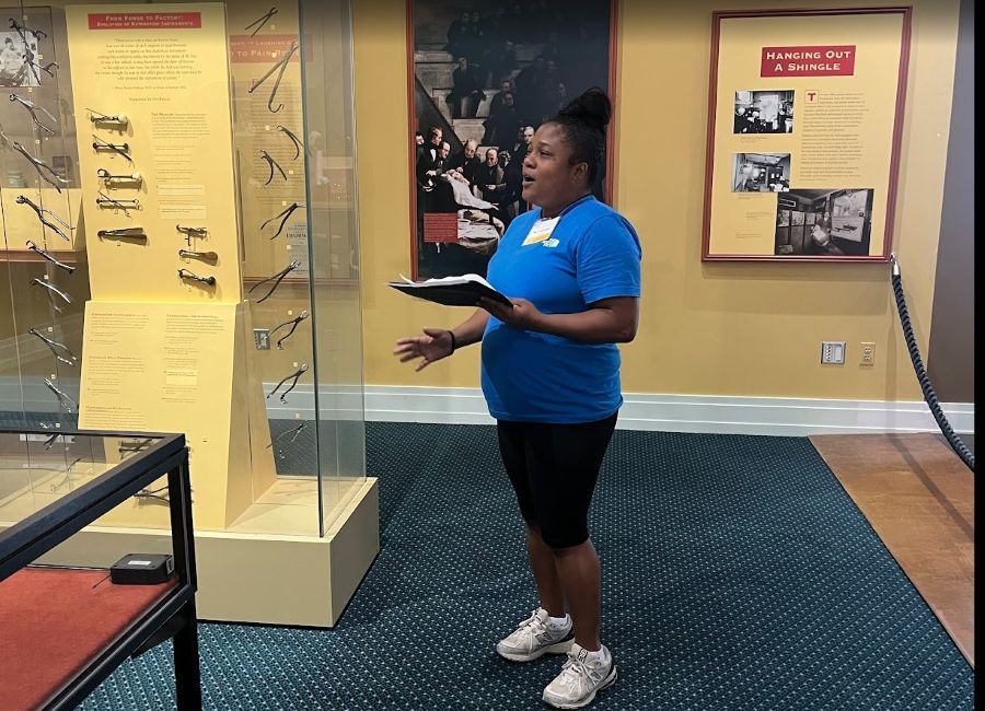 SADE-museum-tour – CareQuest Institute a Woman in a Blue Shirt Speaks or Presents Beside Museum Display Cases, Holding a Notebook and Wearing a Name Badge in a Gallery Setting.