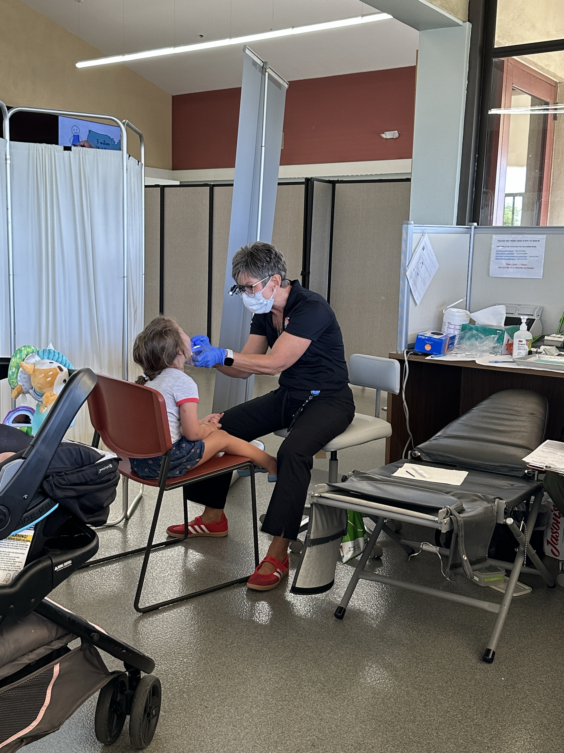 Image 2 – CareQuest Institute Healthcare Worker in a Mask and Gloves Tending to a Child Seated in a Chair Inside a Clinic, with Privacy Screens and Medical Supplies Nearby.