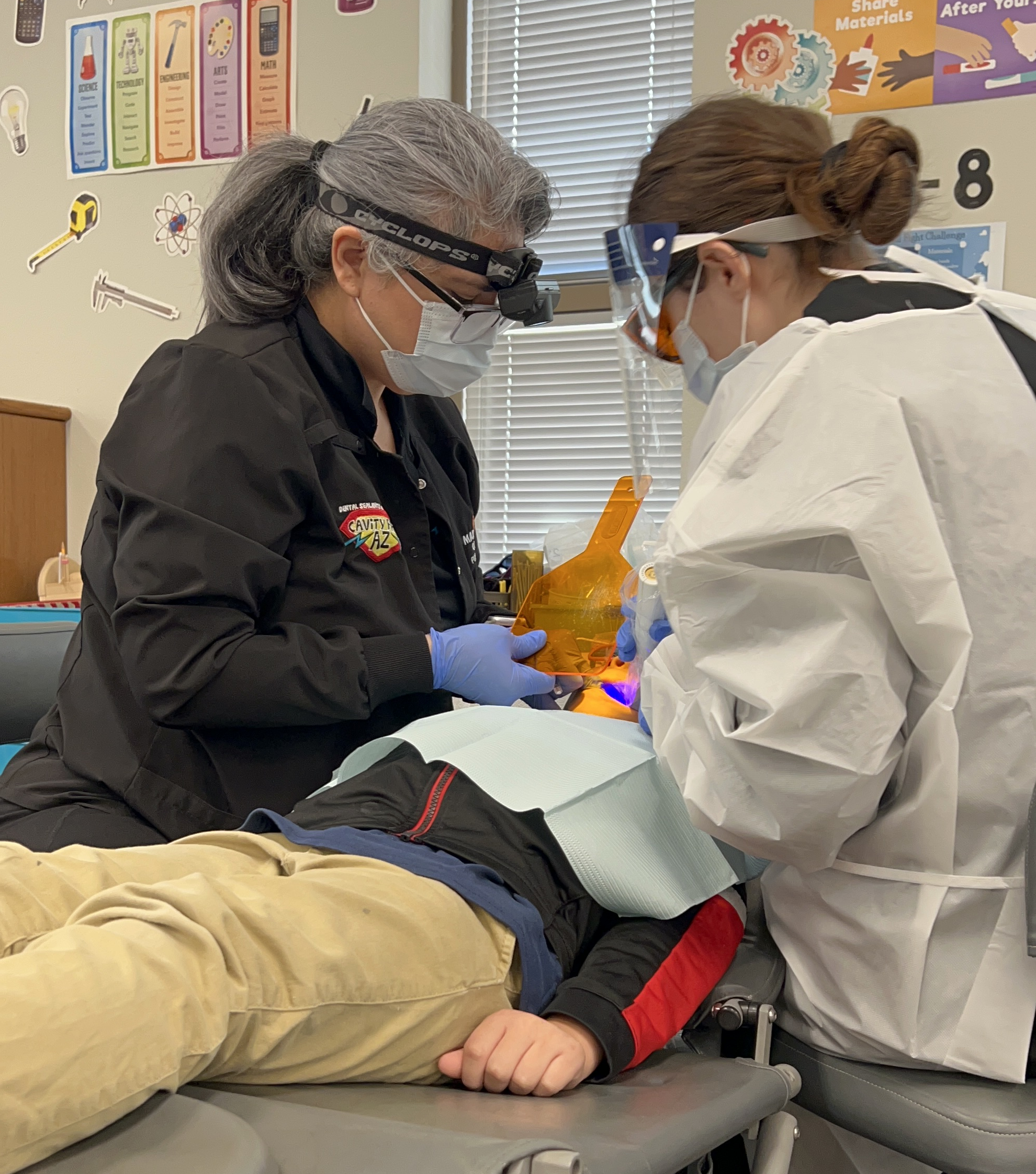 Two Clinicians in Ppe (gloves, Masks, Goggles/face Shield) Perform a Dental Procedure Beside a Patient in a Clinic with a Gray Chair and Orange Instruments Nearby.
