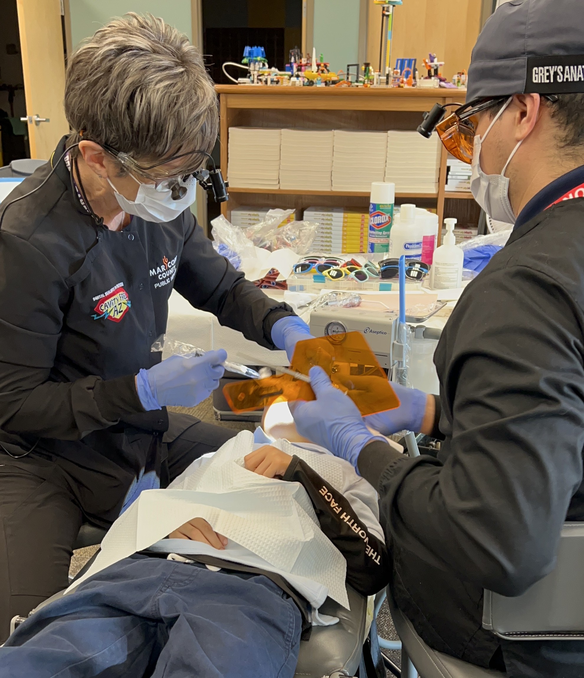 Social Media 2_Cropped – CareQuest Institute Two Clinicians in Masks and Gloves Perform a Dental Procedure, Using Headlamps and Orange Protective Shields over Their Glasses.