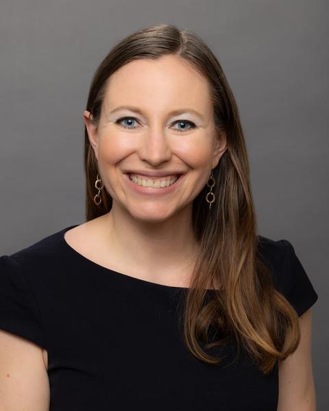 Head-and-shoulders Portrait of a Smiling Woman with Long Light Brown Hair, Wearing a Black Top and Gold Earrings, Against a Gray Backdrop.
