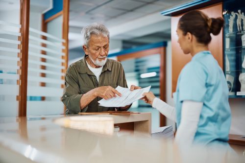 Older Man in a Green Shirt Hands Papers to a Nurse at a Reception Desk in a Clinic Lobby.
